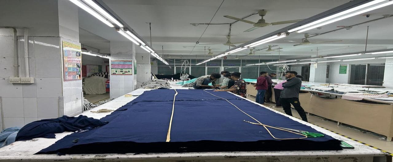 Long cutting table with fabric spread, viewed down its length toward workers marking a pattern